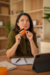 Woman holding an orange credit card and looking thoughtfully in a modern apartment
