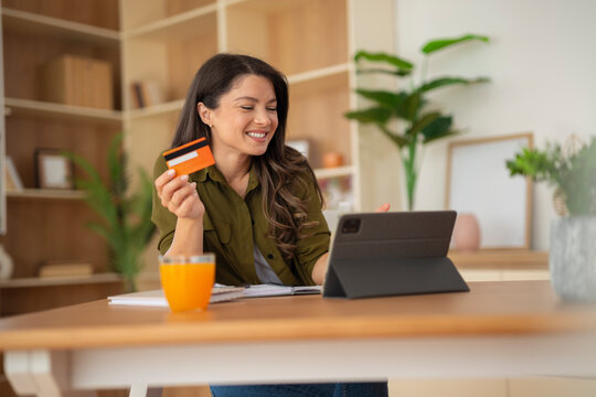 Happy woman holding a credit card while sitting at a kitchen table with a tablet.
