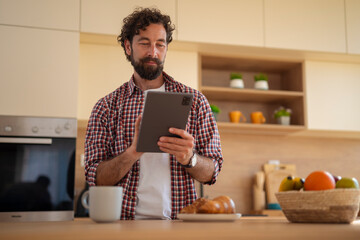 Bearded man focused while using a tablet in a cozy kitchen with coffee and bread.
