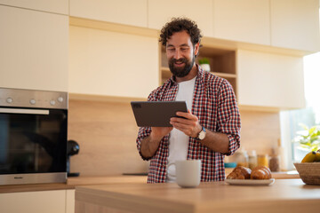 Bearded man smiling while using a tablet in a modern kitchen with a coffee cup nearby.

