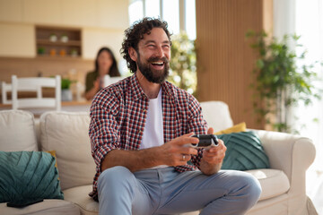 Smiling man holding a video game controller while sitting on a couch with partner in the...