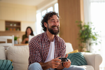 Smiling man holding a video game controller while sitting on a couch with partner in the...