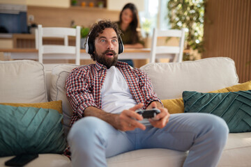Obraz premium Surprised young man playing video games with controller while sitting on couch at home. 