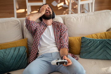 Frustrated young man holding video game controller and resting head on hand while sitting on couch...