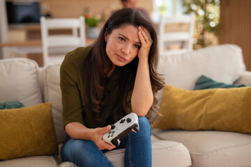 Frustrated young woman holding video game controller and resting head on hand at home.
