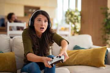 Focused young woman wearing headphones and playing video games with intense expression at home.
