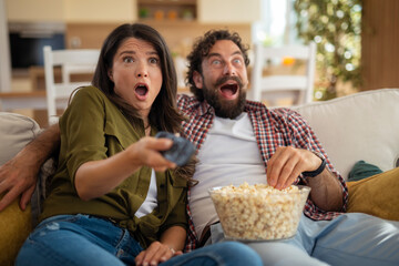 Surprised young couple watching TV and holding remote control while eating popcorn at home.
