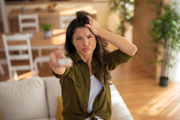 Frustrated woman holding remote control trying to turn on air conditioner that is not working, feeling hot at home.
