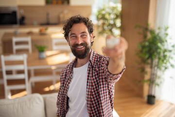 Smiling man holding air conditioner remote control and turning on the AC in kitchen at home.
