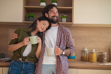 Young couple enjoying morning coffee together in kitchen, woman leaning on man’s shoulder.
