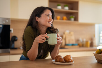 Happy woman holding coffee mug and leaning on kitchen counter while enjoying morning.
