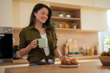 Young woman holding coffee mug and looking at plate of croissants in kitchen at home.

