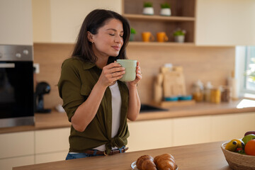 Young woman enjoying aroma of coffee while standing in kitchen with breakfast on table.
