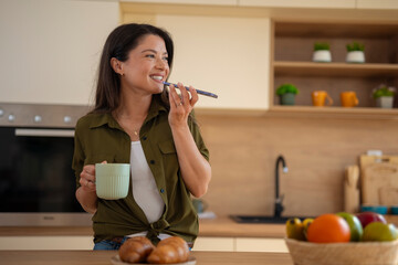 Young woman recording voice message on smartphone while holding coffee mug in kitchen.
