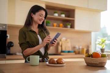 Smiling woman using smartphone in kitchen while enjoying morning coffee and breakfast.
