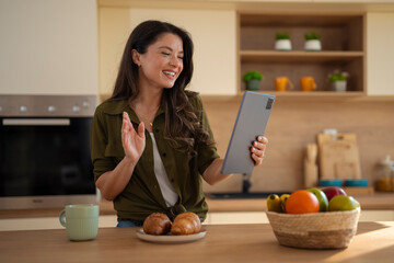 Young woman waving during video call on tablet while standing in kitchen at home.
