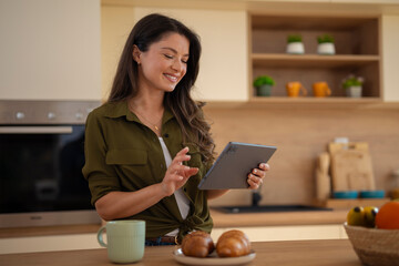 Smiling woman using digital tablet in kitchen, enjoying morning with coffee and breakfast.
