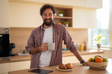 Young man holding coffee mug in kitchen, smiling and enjoying morning at home.
