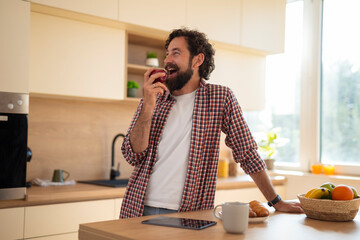 Smiling man eating apple while standing in kitchen at home, enjoying morning snack.
