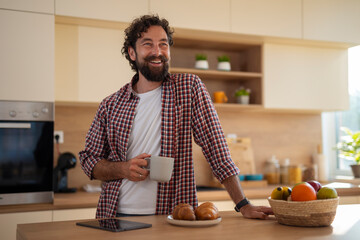 Happy man standing in kitchen, holding coffee mug and enjoying morning at home.
