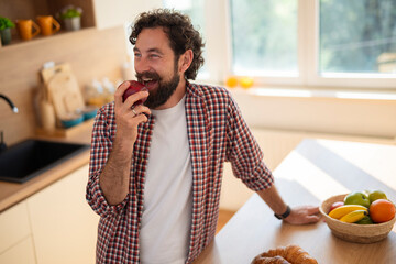 Smiling man eating apple in kitchen at home, enjoying healthy snack in casual setting.
