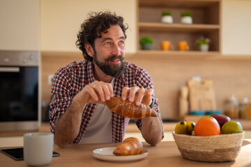 Young man sitting in kitchen, enjoying breakfast with croissant and coffee in modern home.
