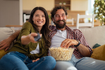 Happy young couple sitting on the couch watching TV and eating popcorn together at home. Relaxed atmosphere and quality time in a modern living room.
