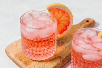 Two glasses of a pink lemonade cocktail with ice and a grapefruit slice garnish on a wooden board, set against a light background. The drink is refreshing and suitable for a summer setting.