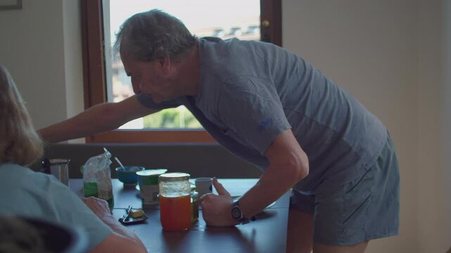Senior man preparing casual breakfast at kitchen table by window, reaching for bottle and pouring milk or yogurt into bowl, surrounded by food jars and cups
