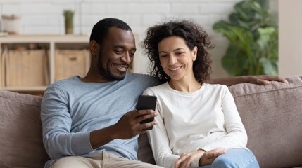Mid adult man relaxing on sofa and showing new app to african american wife on cellphone. Middle eastern man and woman sitting on couch at home and using mobile phone to do a video call with family.,