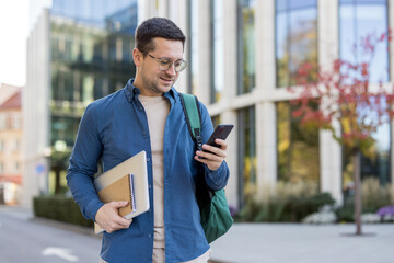 A young man in glasses is using his phone outside a modern building, carrying a laptop and notebook.