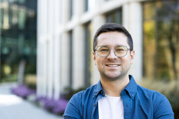 Smiling man in glasses poses in front of a modern building. The photo captures a friendly...