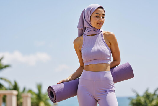 Young woman wearing hijab holding yoga mat outdoors on sunny day, expressing fitness enthusiasm and relaxation in lavender sportswear - Powered by Adobe