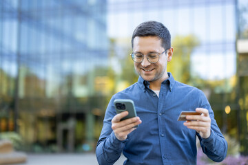 A man holding a credit card and smartphone smiles in front of a modern building.