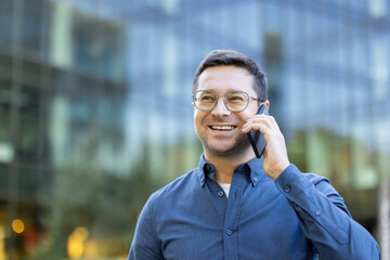 A smiling man with glasses talks on his phone in front of a modern building. Business call.