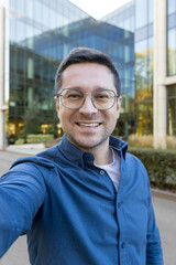 A man wearing glasses takes a selfie in front of a modern glass building, smiling towards the...