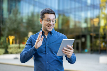 A man in eyeglasses waves during a video call while holding a tablet outdoors in front of a blurred...