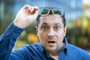 Man wearing glasses in a state of surprise and shock. The background is blurred with natural light.