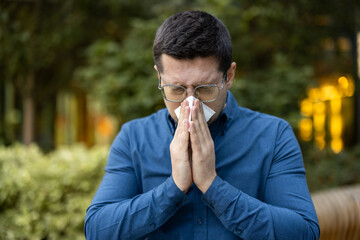 A man in glasses outside blows his nose with a tissue, likely feeling unwell, amid greenery.