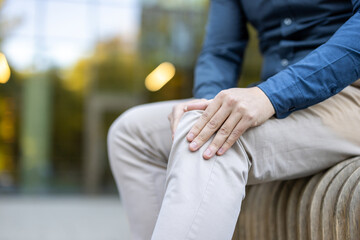A man, dressed in a blue shirt and beige pants, sits with his hands on his knee, possibly...