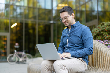 A man in a blue shirt works on a laptop outside, wearing glasses and earphones, smiling. A bicycle...