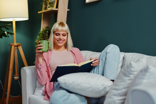 Smiling woman reading a book and drinking tea on sofa at home