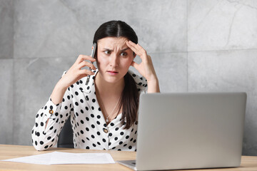 Young businesswoman with papers and laptop complaining to someone via phone at wooden table indoors