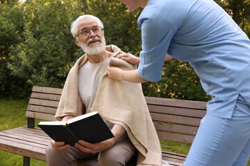 Fototapeta premium Nurse supporting smiling elderly man in glasses with book outdoors, closeup