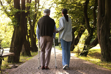 Caregiver supporting elderly man who using walking frame outdoors, back view