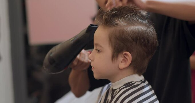 Hairdresser Drying Boy s Hair with Hair Dryer in Beauty Salon. Professional stylist gently dries a young boy s hair after a fresh haircut. Modern men's hairstyle for children in a cozy beauty salon.