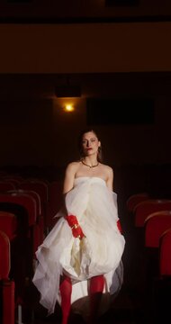 A glamorous woman in a white dress walks down the aisle between the seats in the theater auditorium. She looks at the camera.