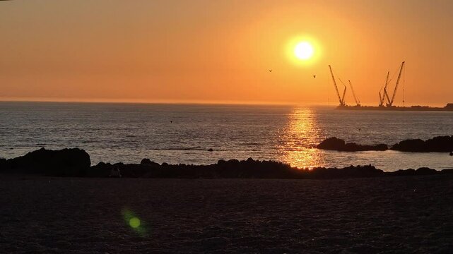 Sunset at Homem do Leme beach in Porto, Portugal. A quiet moment by the Atlantic as the golden sun reflects on the water, silhouetting rocks, cranes.