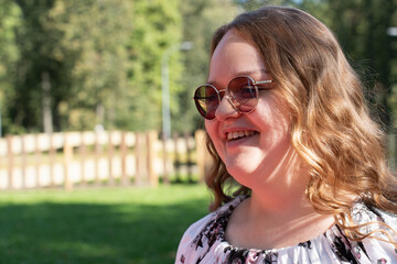 Smiling young woman with long curly hair and sunglasses standing outdoors on a sunny day, looking at the camera.