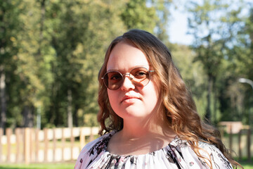 Young woman with long curly hair and sunglasses standing outdoors on a sunny day, looking at the camera.